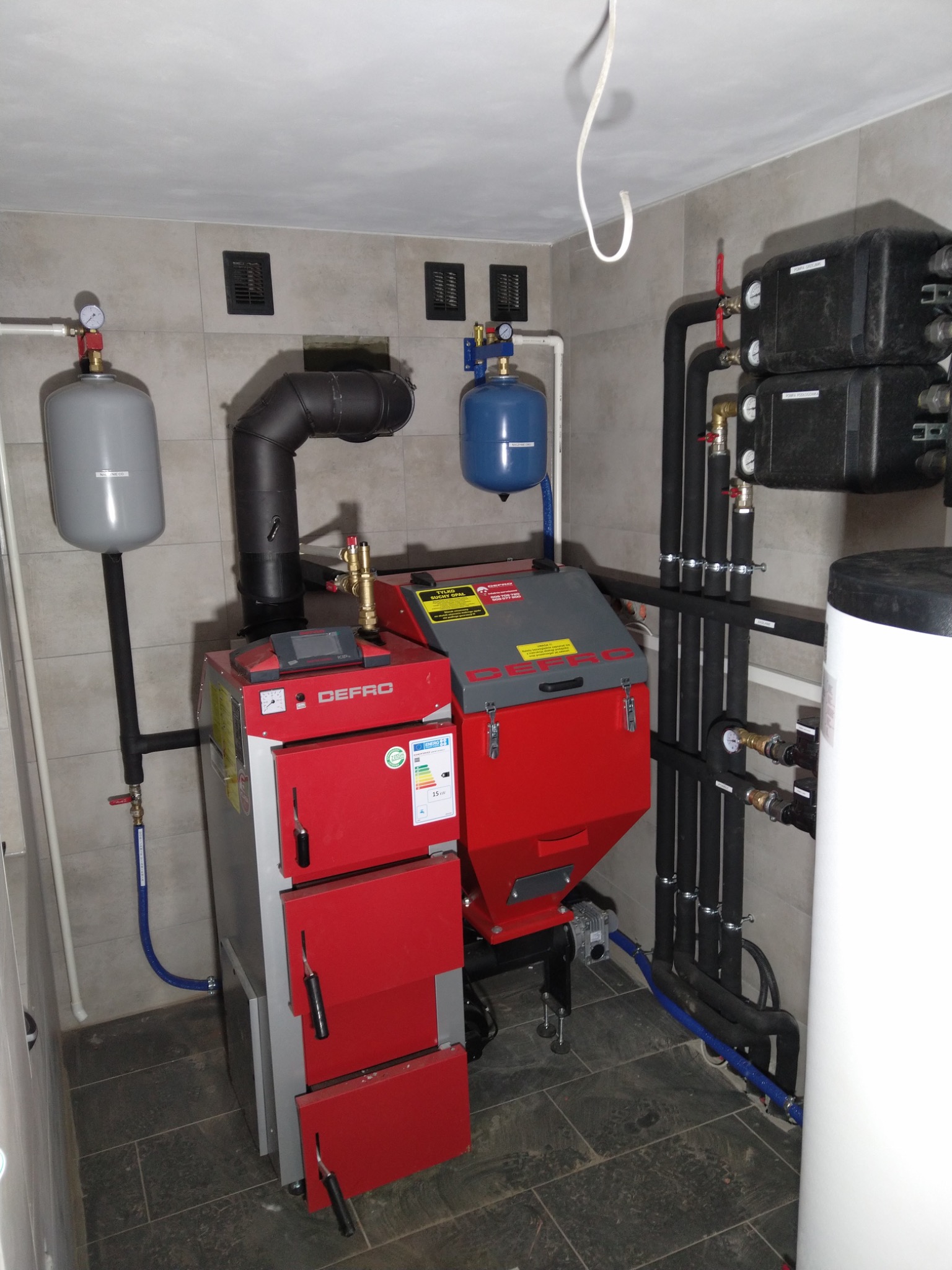 Nowoczesny, automatyczny piec na paliwo stałe Defro, z czerwonymi drzwiczkami i szarym korpusem, zainstalowany w kotłowni z szarymi płytkami, widoczne czarne rury instalacji grzewczej, zbiorniki...