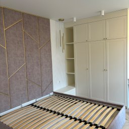 Iryna Zhabenko - Sypialnia z łóżkiem bez materaca, tapicerowanym wezgłowiem w geometryczne wzory z złotymi ramkami i kremową szafą wnękową z półkami.