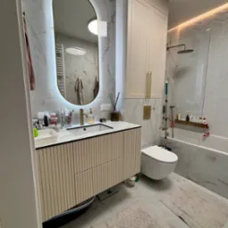 Nowoczesna łazienka z umywalką na szafce w paski, owalnym lustrem z podświetleniem LED i białymi płytkami imitującymi marmur. Widoczna toaleta i wanna z parawanem.