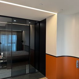 Iryna Zhabenko - Nowoczesna szafa przesuwna z czarnymi ramami i lustrami w sypialni. Widoczne oświetlenie LED i fragment szafy z pomarańczowo-białymi panelami.