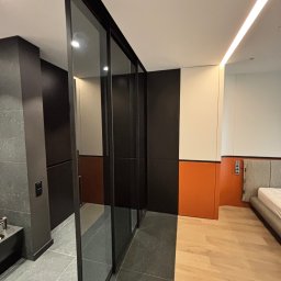 Iryna Zhabenko - Nowoczesna szafa przesuwna z czarnymi ramami i szklanymi drzwiami, oddzielająca łazienkę od sypialni we Wrocławiu. Minimalistyczny design, widoczne różne wykończenia podłóg.