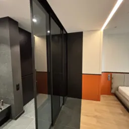 Nowoczesna szafa przesuwna z czarnymi ramami i szklanymi drzwiami, oddzielająca łazienkę od sypialni we Wrocławiu. Minimalistyczny design, widoczne różne wykończenia podłóg.