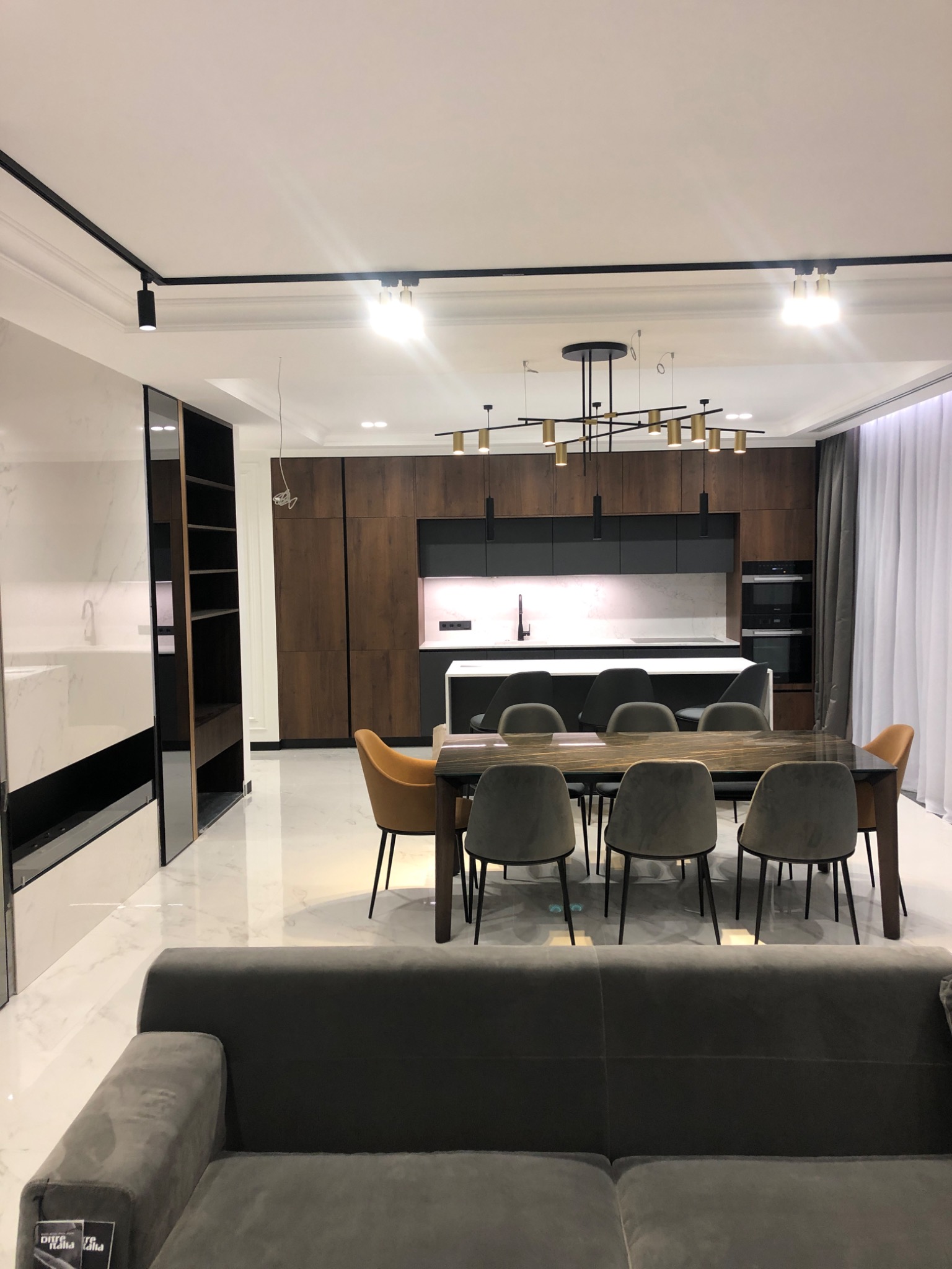 Nowoczesne wnętrze z otwartą kuchnią: drewniane fronty szafek, biały blat wyspy kuchennej, marmurowa ściana, stół z krzesłami w odcieniach szarości i musztardy, oświetlenie punktowe i designerski...