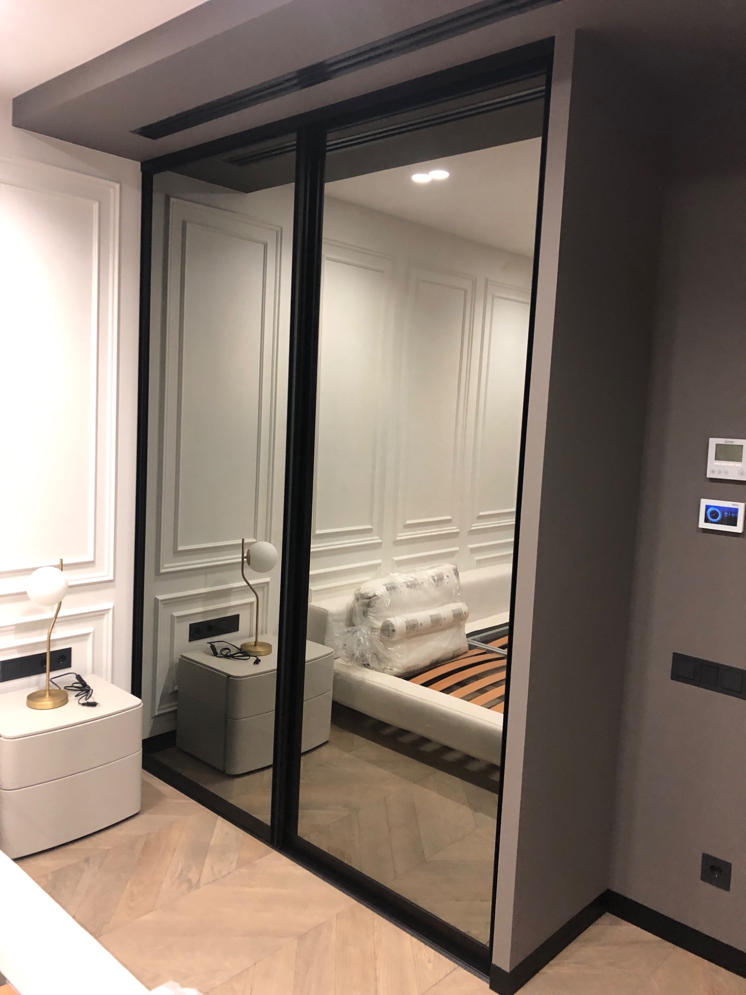 Nowoczesna szafa wnękowa z przesuwnymi drzwiami lustrzanymi, odbijająca fragment sypialni z łóżkiem i stolikami nocnymi na parkiecie w jodełkę.