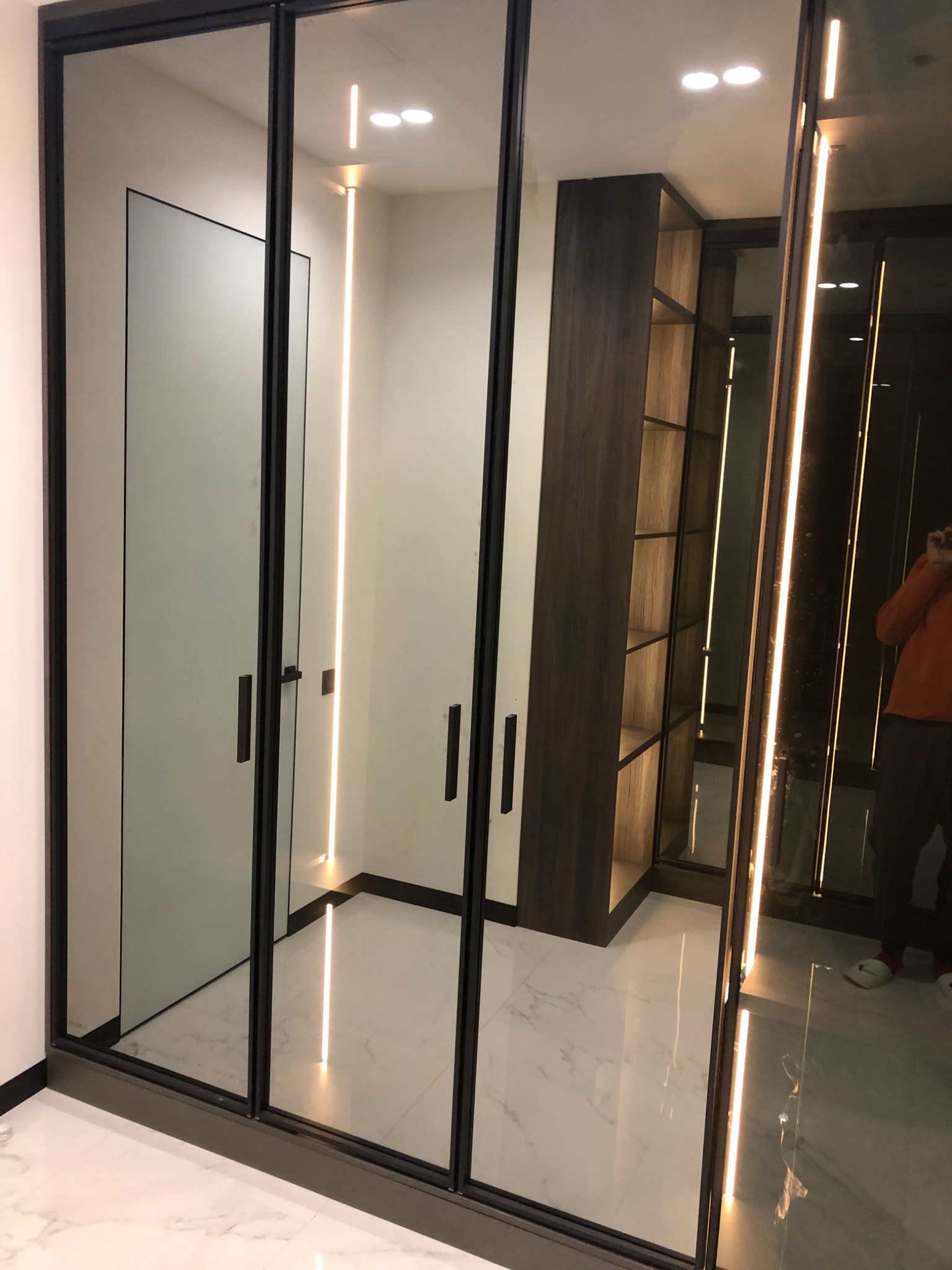 Nowoczesna szafa wnękowa z przesuwnymi drzwiami, czarne ramy, podświetlenie LED wewnątrz, marmurowa podłoga, widoczny fragment garderoby z półkami.