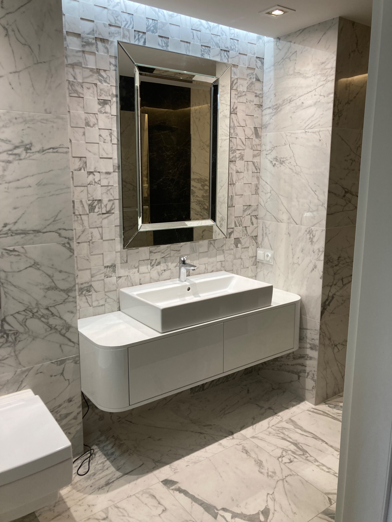 Elegancka łazienka z marmurowymi płytkami, wiszącą białą szafką z umywalką i lustrem w ozdobnej ramie.