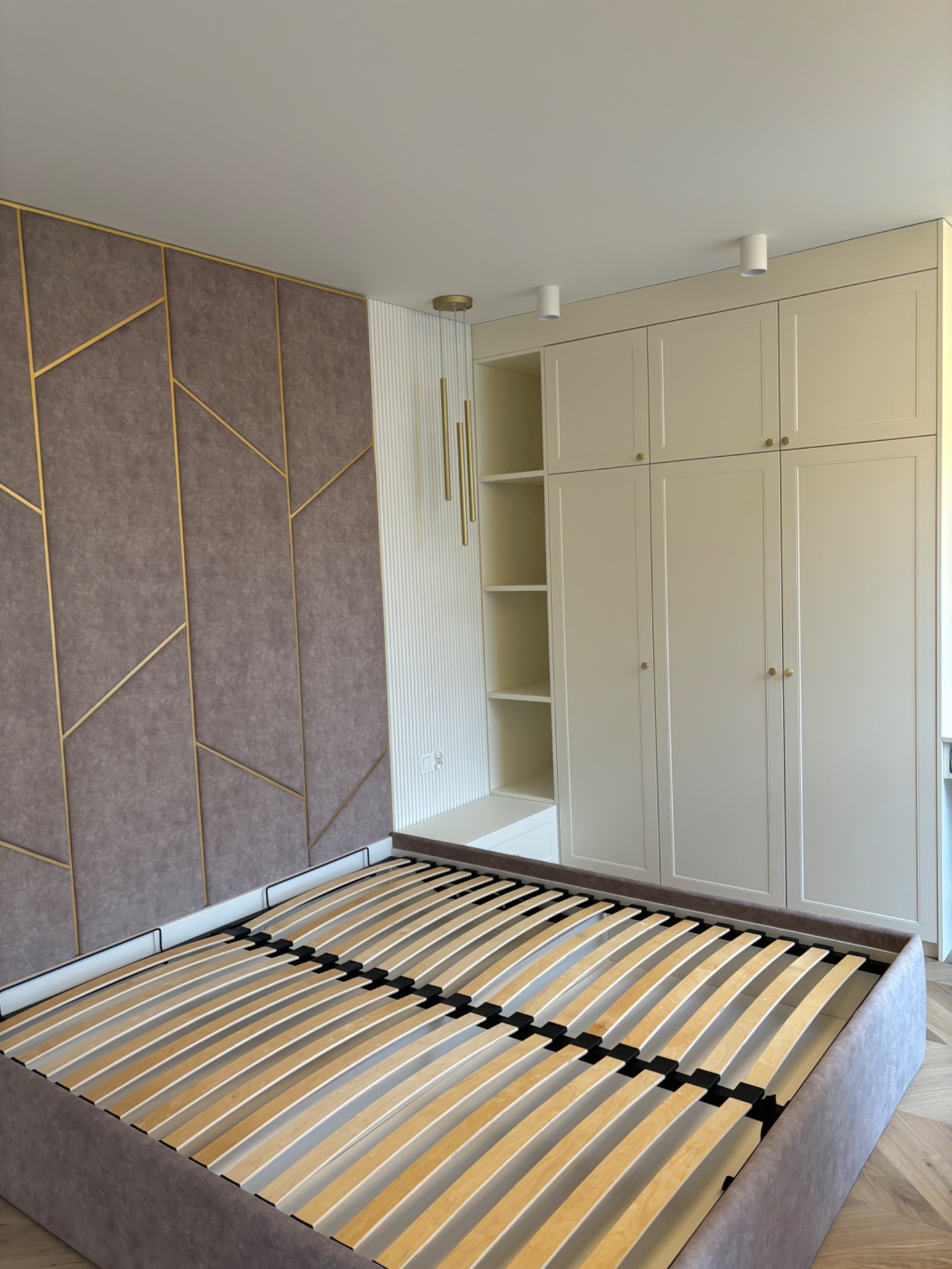 Sypialnia z łóżkiem bez materaca, tapicerowanym wezgłowiem w geometryczne wzory z złotymi ramkami i kremową szafą wnękową z półkami.