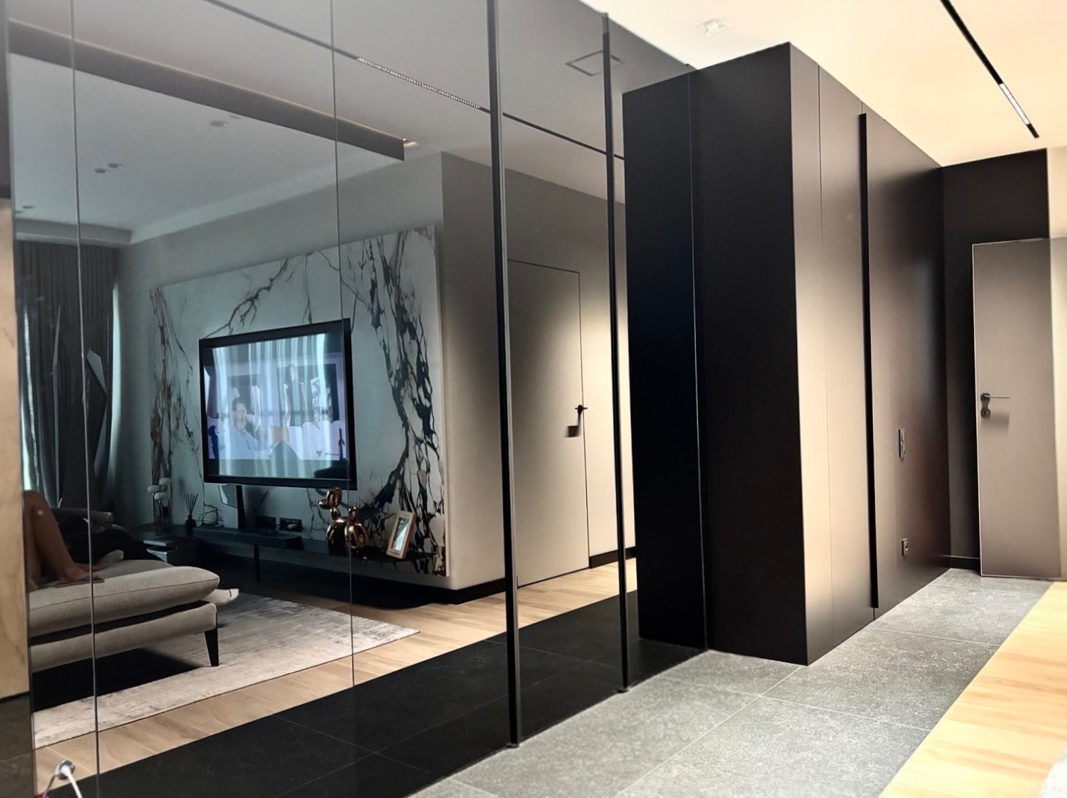 Nowoczesne wnętrze z szafą na wymiar w kolorze czarnym, odbijającą w lustrzanych drzwiach salon z marmurową ścianą i telewizorem. Minimalistyczny design.