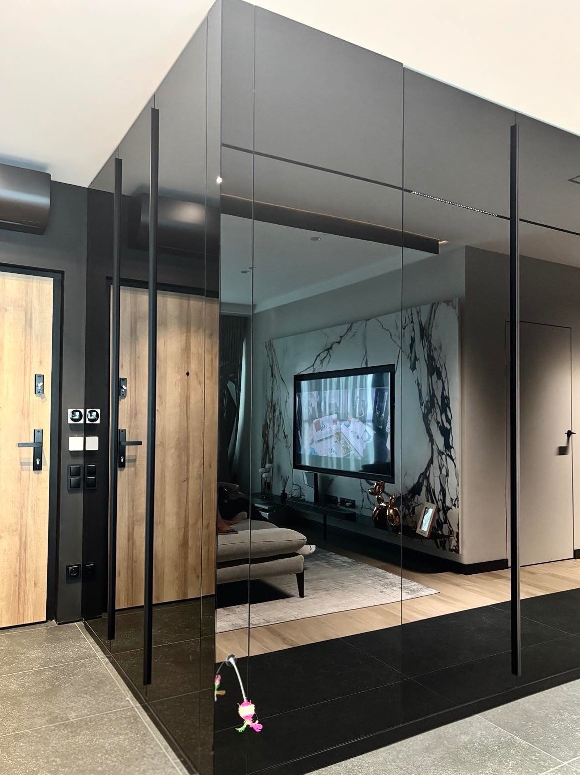 Nowoczesna szafa z lustrzanymi drzwiami przesuwnymi w przedpokoju. Widoczne drzwi wejściowe i fragment salonu z telewizorem na ścianie z marmurowym wzorem.