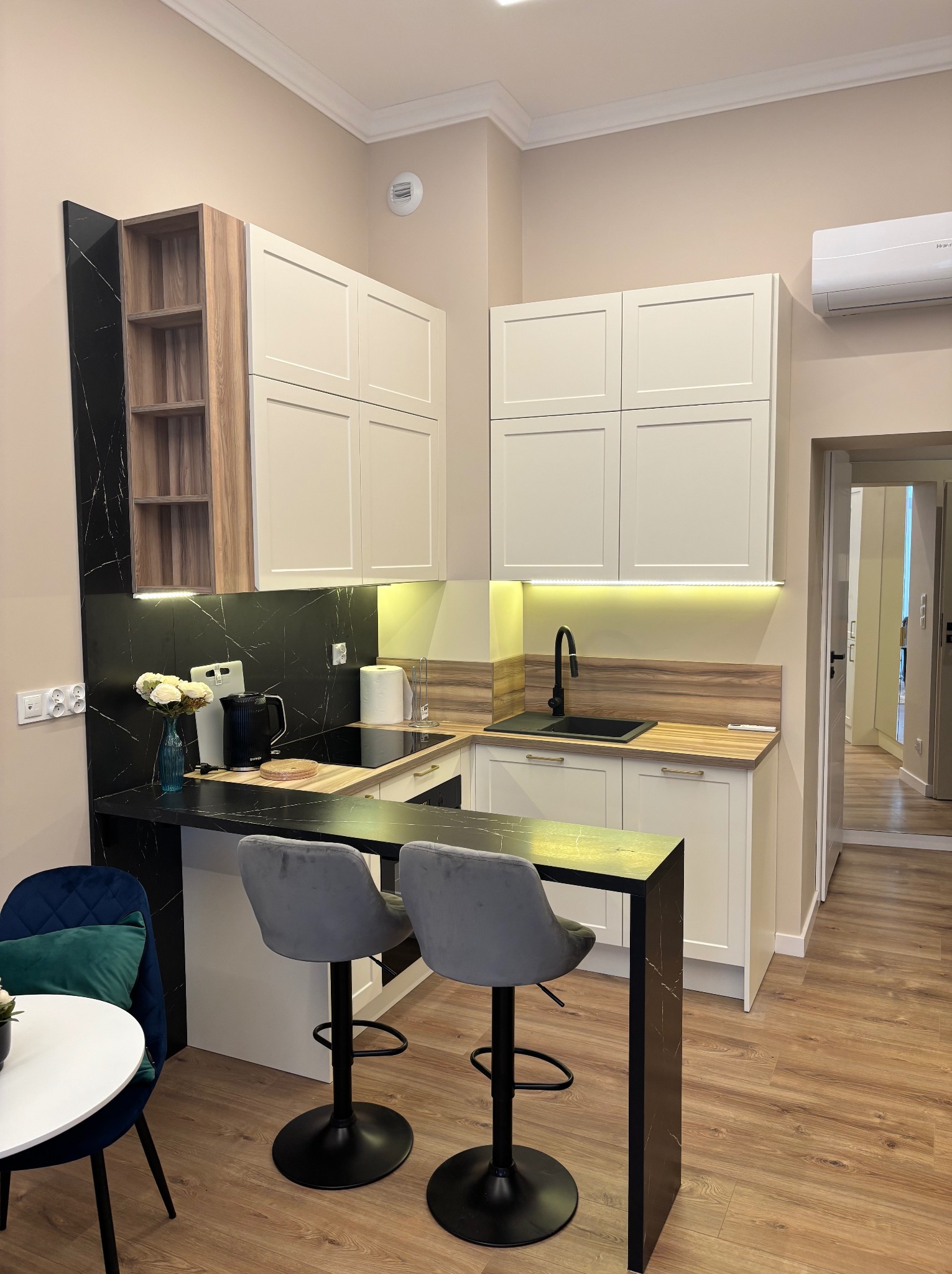 Nowoczesna kuchnia z białymi szafkami, czarnym blatem z marmurowym wzorem i hokerami. Oświetlenie LED pod szafkami górnymi. Widoczne akcesoria kuchenne i kwiaty.