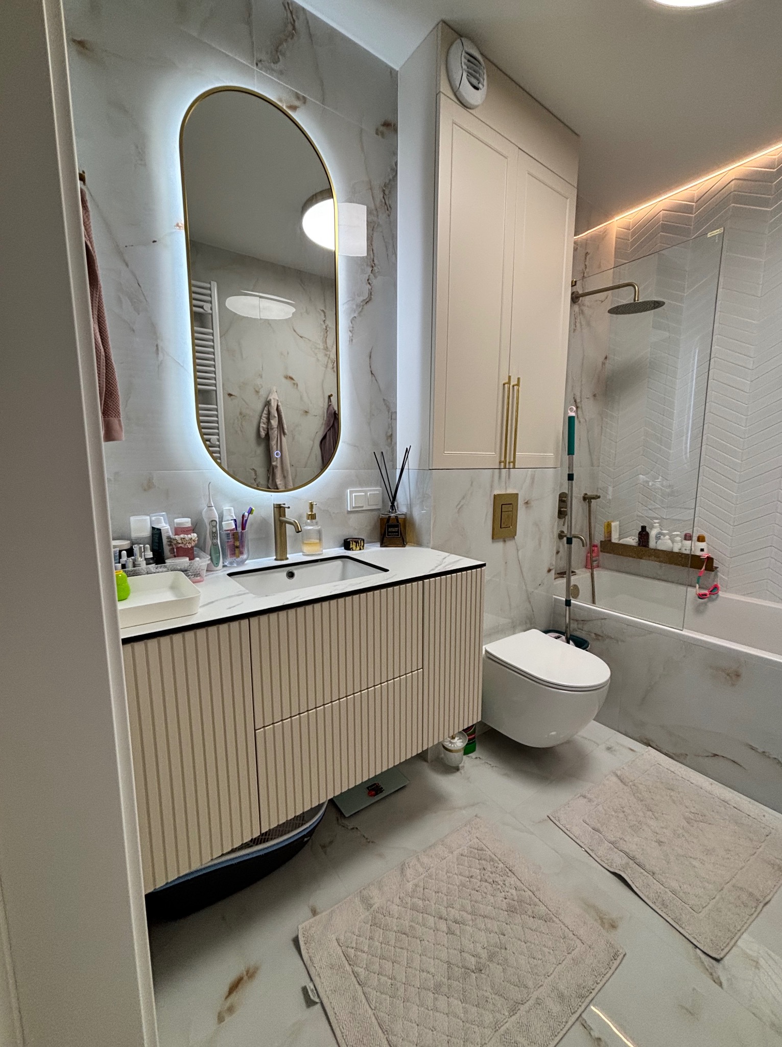 Nowoczesna łazienka z umywalką na szafce w paski, owalnym lustrem z podświetleniem LED i białymi płytkami imitującymi marmur. Widoczna toaleta i wanna z parawanem.