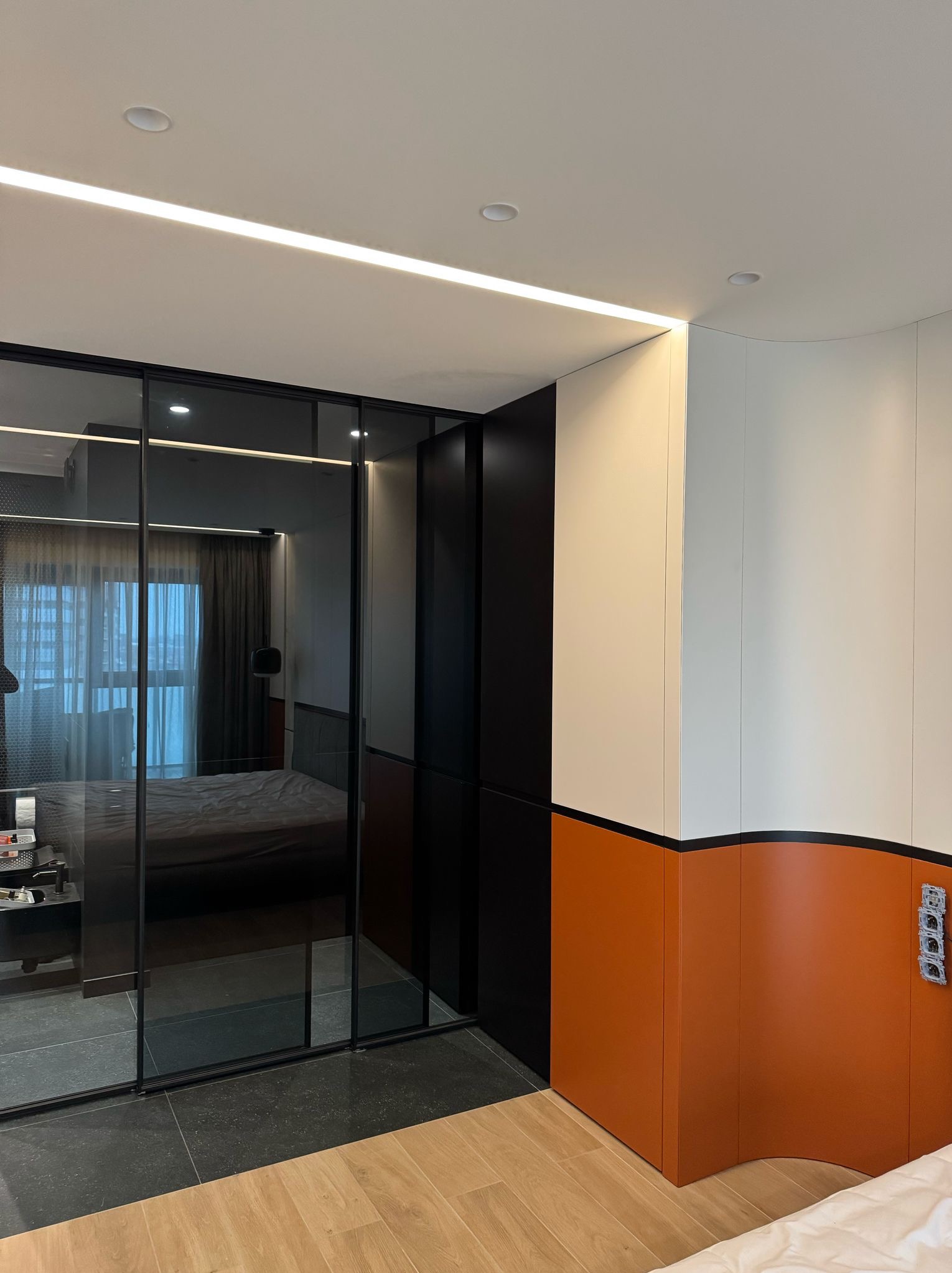 Nowoczesna szafa przesuwna z czarnymi ramami i lustrami w sypialni. Widoczne oświetlenie LED i fragment szafy z pomarańczowo-białymi panelami.