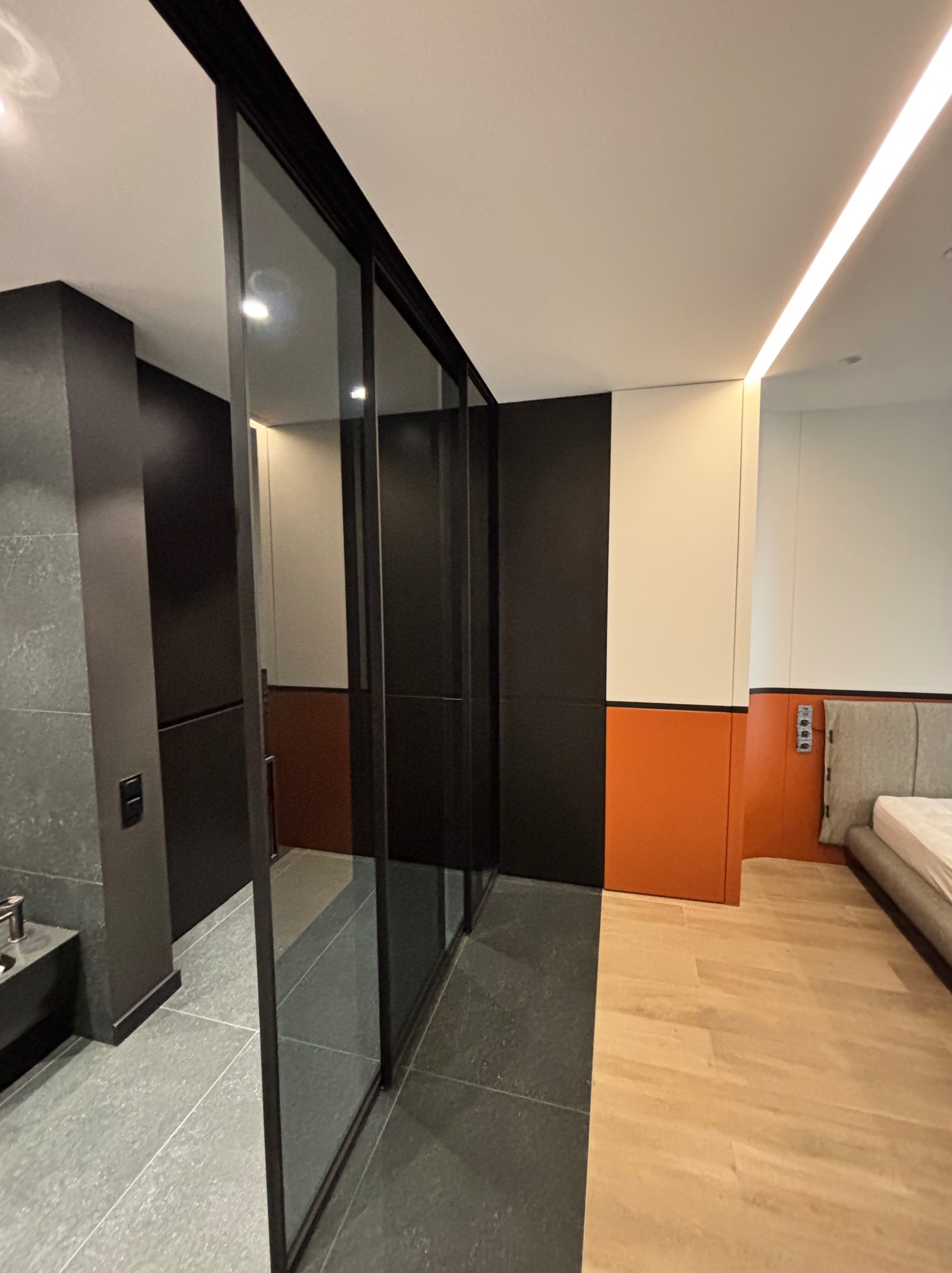 Nowoczesna szafa przesuwna z czarnymi ramami i szklanymi drzwiami, oddzielająca łazienkę od sypialni we Wrocławiu. Minimalistyczny design, widoczne różne wykończenia podłóg.