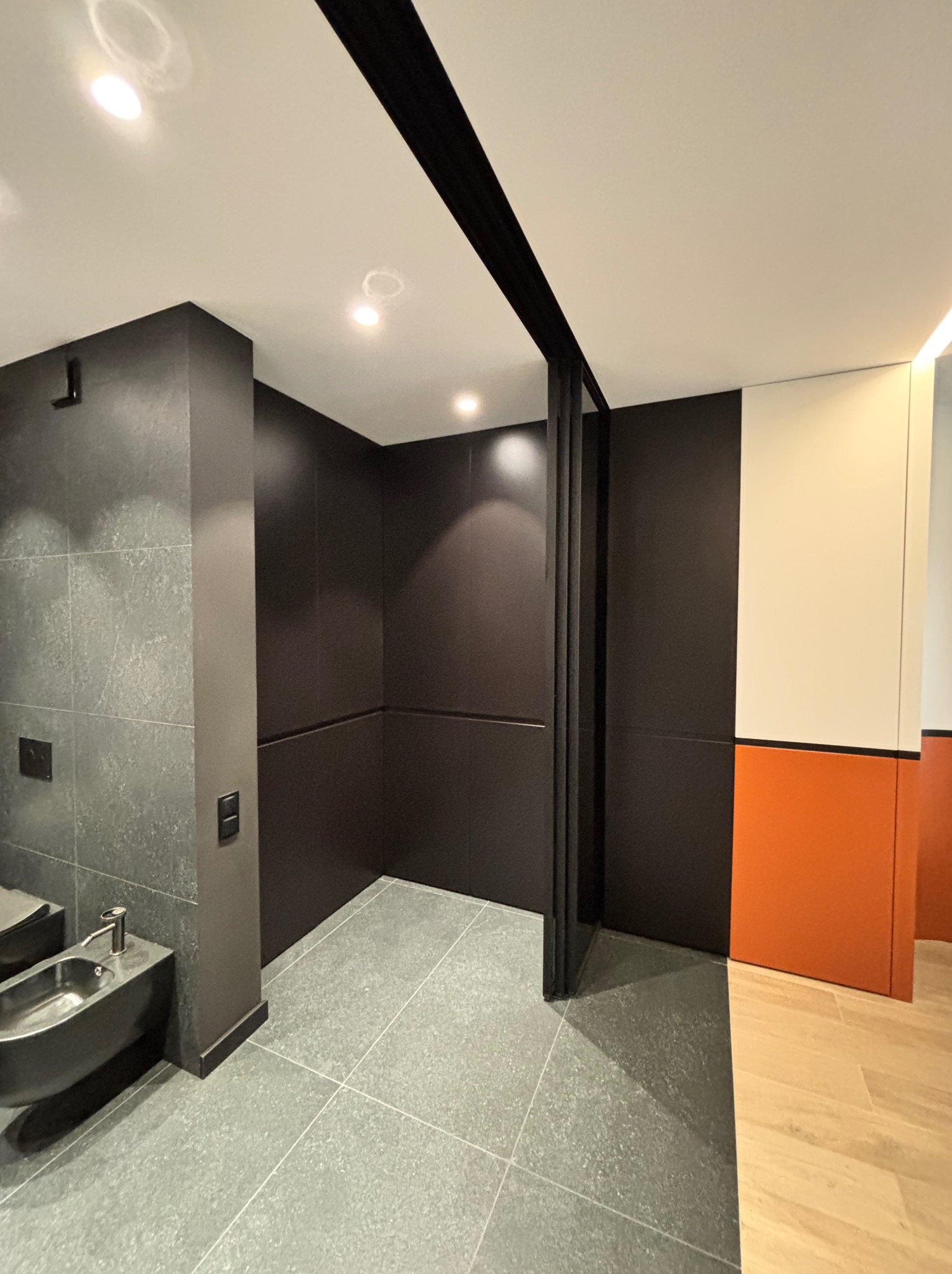 Nowoczesna łazienka z bidetem, szarą płytką i czarnymi elementami: ściany, drzwi przesuwne. Kontrastujące pomarańczowe i białe akcenty na ścianie.