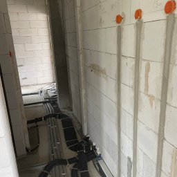 AROS Adam Rosiek - Wnętrze w trakcie prac: widoczne kable elektryczne prowadzone wzdłuż ściany z betonu komórkowego, z pomarańczowymi puszkami instalacyjnymi. Etap przed tynkowaniem.