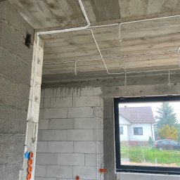AROS Adam Rosiek - Surowy stan: białe kable elektryczne prowadzone po suficie i ścianach z betonu komórkowego w nowym budynku. Pomarańczowe puszki elektryczne w ścianach.