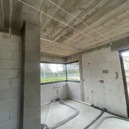 AROS Adam Rosiek - Surowy stan wnętrza z widoczną instalacją elektryczną i hydrauliczną. Kable i rury prowadzone po suficie i podłodze w nowym budynku z oknem na zewnątrz.