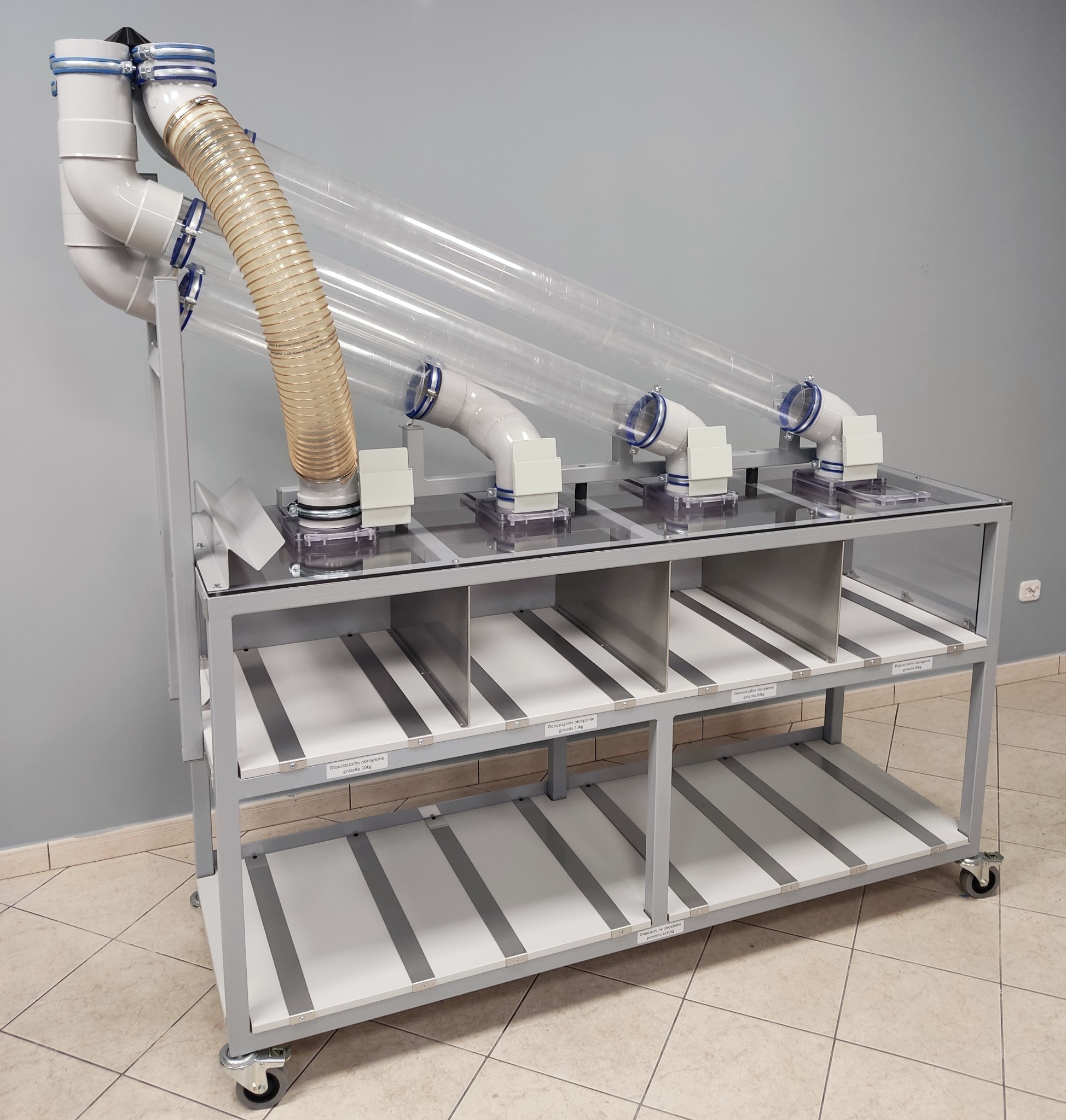 Stalowa konstrukcja z przezroczystymi rurami i elastycznym przewodem wentylacyjnym, prawdopodobnie element zautomatyzowanego stanowiska montażowego do segregacji detali, na kółkach, z półkami...