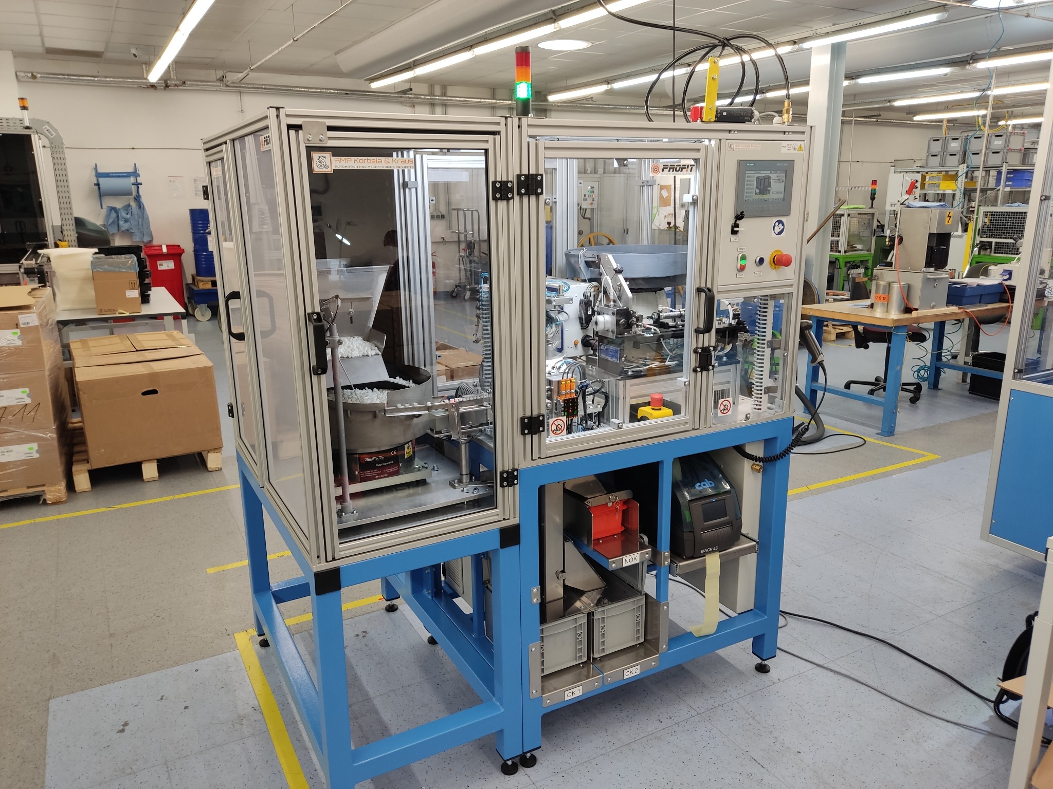 Automatyczna maszyna do montażu o-ringów z przezroczystymi osłonami, podświetlonym wnętrzem, panelem sterowania i podajnikiem wibracyjnym, umieszczona w hali produkcyjnej.