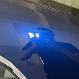 Błyszczący, granatowy lakier samochodu po polerowaniu, z widocznym odbiciem osoby robiącej zdjęcie; fragment okna zabezpieczony taśmą malarską.