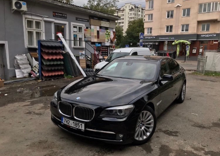 Czarna limuzyna BMW serii 7 zaparkowana przed sklepem z materiałami budowlanymi, w tle budynki mieszkalne.
