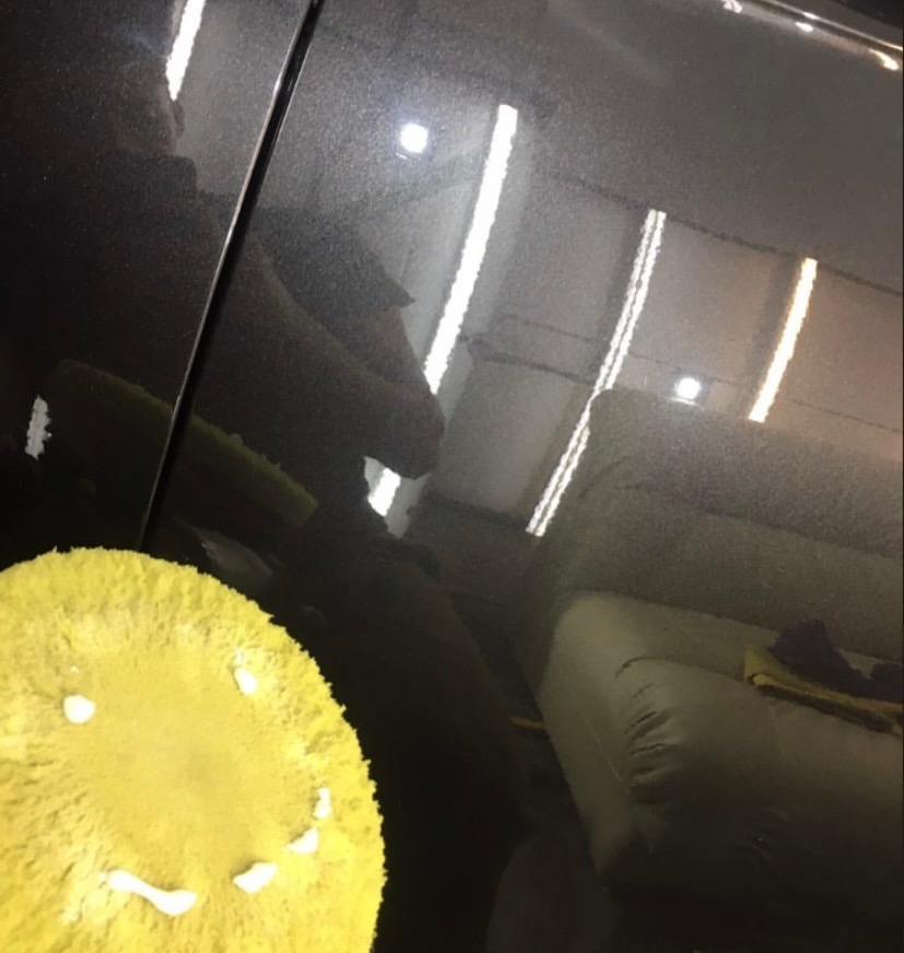 Polerowanie lakieru ciemnego samochodu żółtą gąbką z resztkami pasty polerskiej, widoczne odbicia lamp warsztatowych.