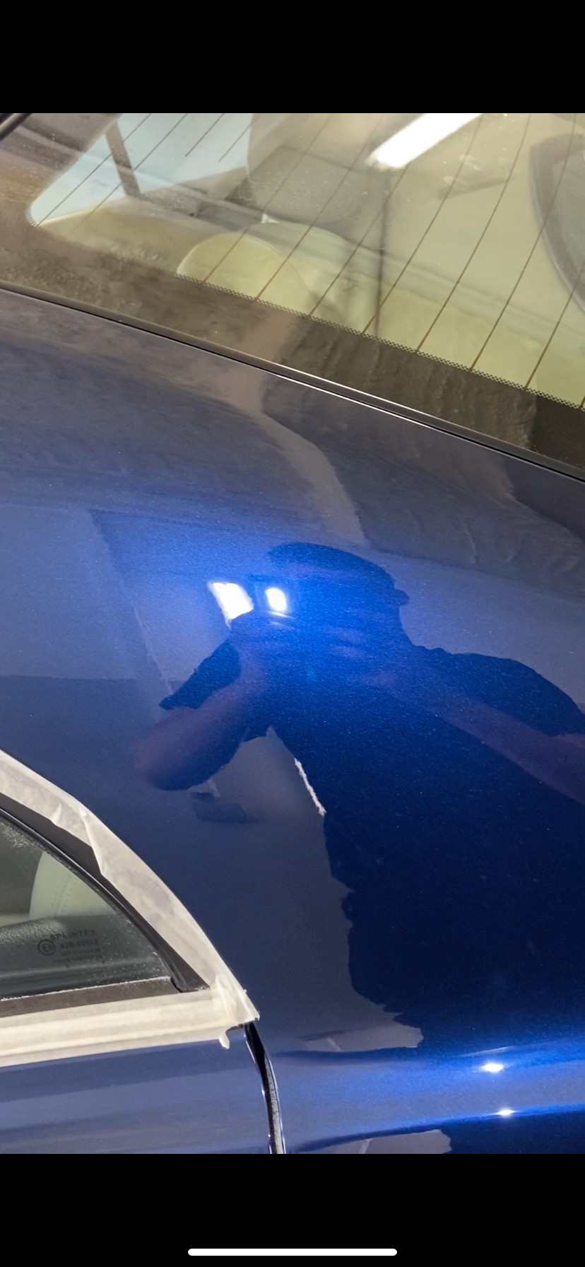 Błyszczący, granatowy lakier samochodu po polerowaniu, z widocznym odbiciem osoby robiącej zdjęcie; fragment okna zabezpieczony taśmą malarską.