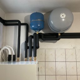KWADRAT MB ŻABKIEWICZ ANETA - Instalacja grzewcza z dwoma zbiornikami wyrównawczymi (niebieski i szary) oraz izolowanymi rurami, zamontowana na tle ściany wyłożonej beżowymi płytkami.