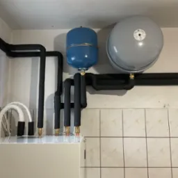 Instalacja grzewcza z dwoma zbiornikami wyr&oacute;wnawczymi (niebieski i szary) oraz izolowanymi rurami, zamontowana na tle ściany wyłożonej beżowymi płytkami.