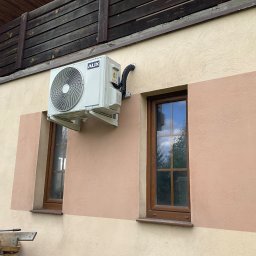 KWADRAT MB ŻABKIEWICZ ANETA - Zewnętrzna jednostka klimatyzacji AUX zamontowana na ścianie domu z dwoma oknami; widoczne przewody i rury odprowadzające.