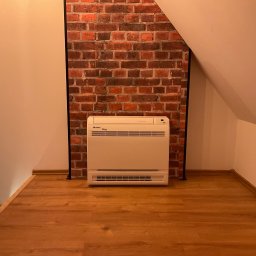 KWADRAT MB ŻABKIEWICZ ANETA - Klimatyzator podłogowy marki Gree ustawiony na drewnianej podłodze na poddaszu, z dekoracyjną ścianą z czerwonej cegły w tle.
