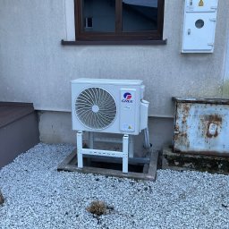 KWADRAT MB ŻABKIEWICZ ANETA - Zewnętrzna jednostka klimatyzacji marki Gree, zamontowana na białej metalowej konstrukcji na żwirowym podłożu, obok skorodowanej skrzynki elektrycznej, na tle elewacji domu z oknem.