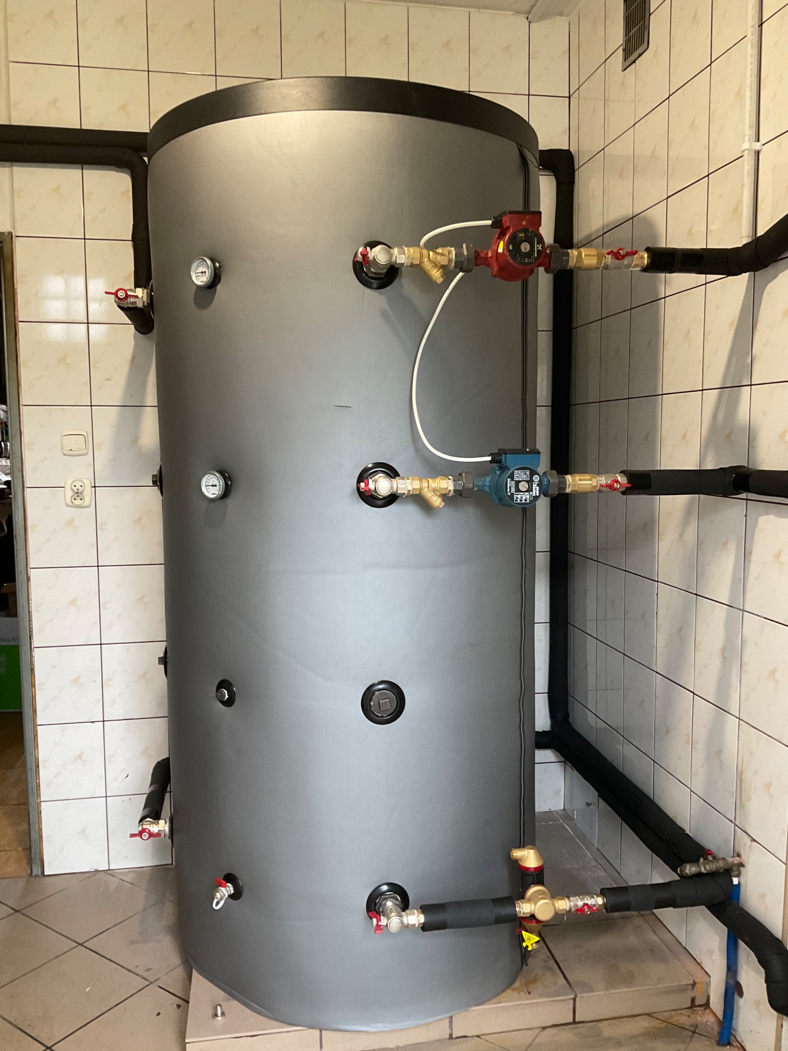 Szary, pionowy bufor ciepła z podłączonymi rurami, zaworami i pompami obiegowymi w pomieszczeniu z beżowymi, kwadratowymi płytkami.