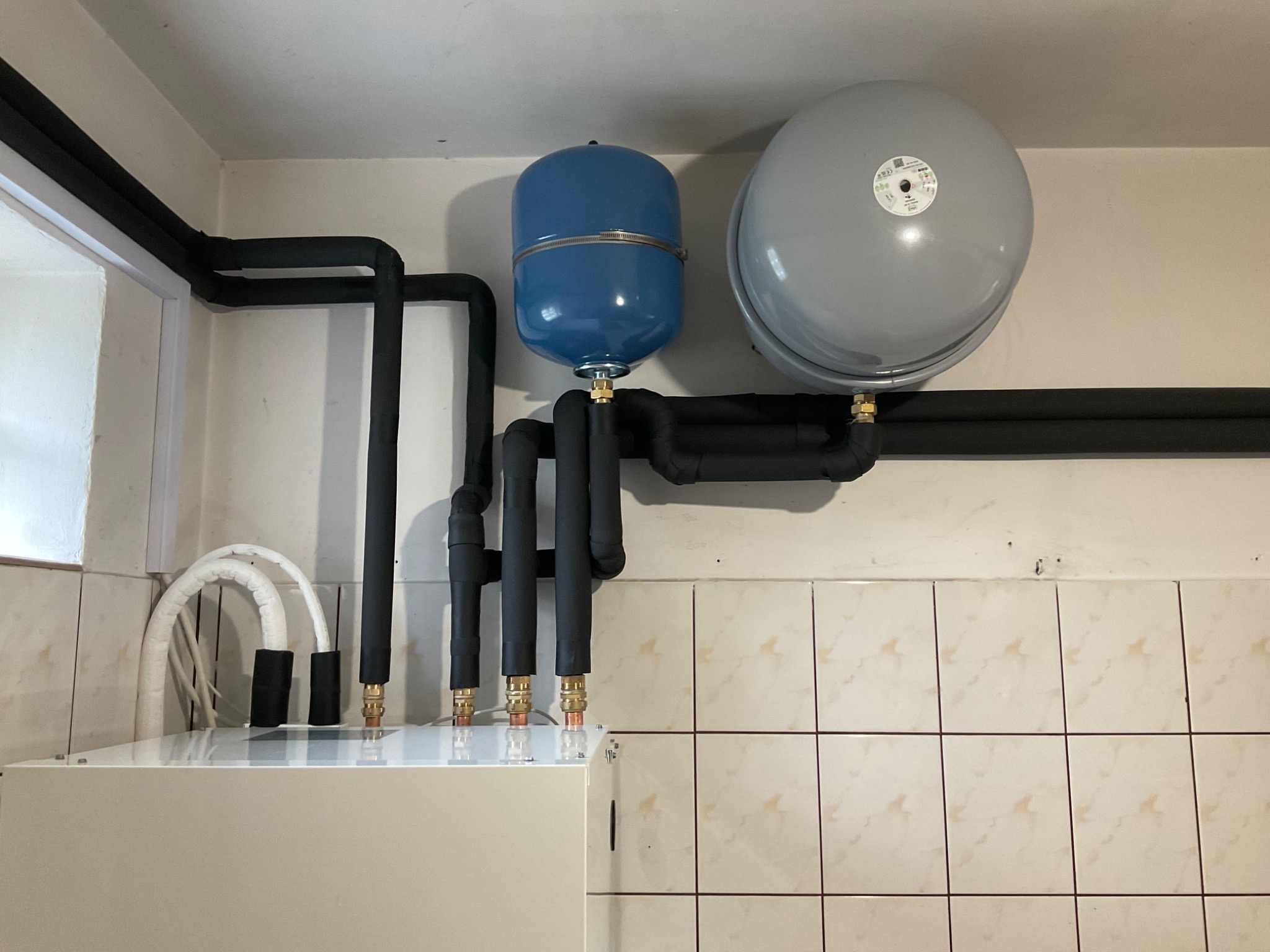 Instalacja grzewcza z dwoma zbiornikami wyrównawczymi (niebieski i szary) oraz izolowanymi rurami, zamontowana na tle ściany wyłożonej beżowymi płytkami.