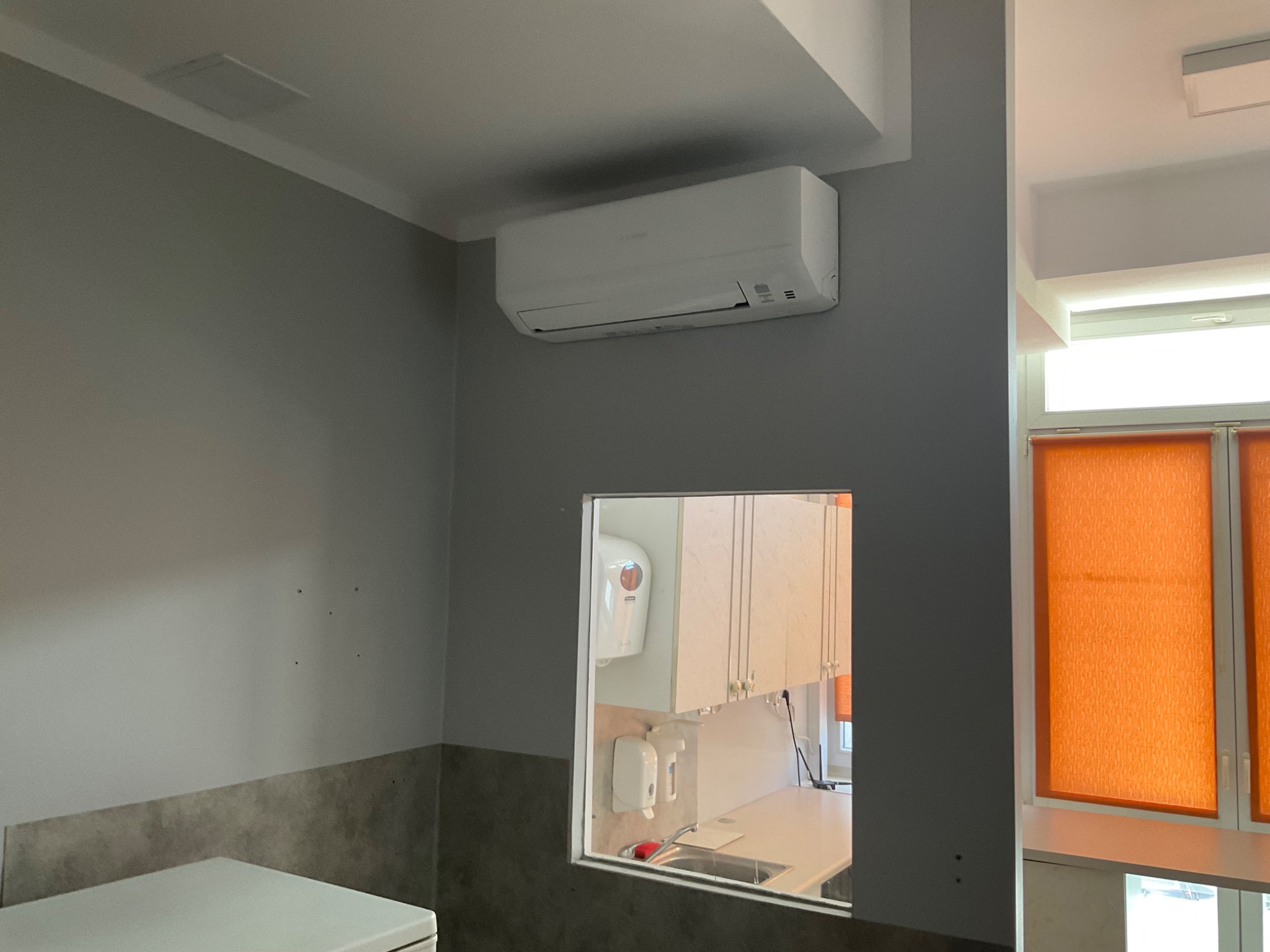 Biała jednostka klimatyzacyjna zamontowana na szarej ścianie, widok przez otwór w ścianie na pomieszczenie z szafkami i zlewem, pomarańczowe rolety w oknie.