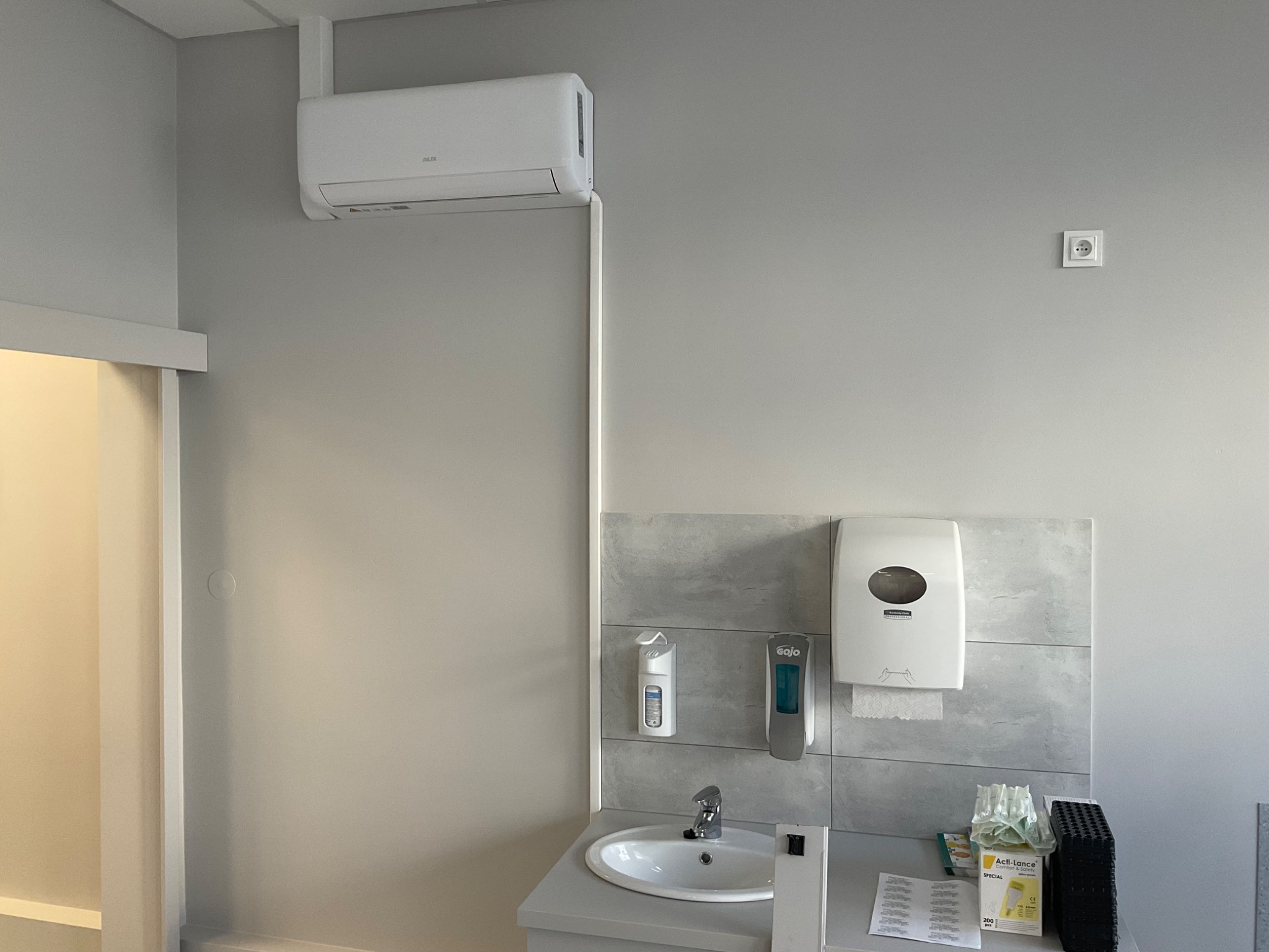 Wnętrze pomieszczenia z zamontowaną białą jednostką klimatyzacji typu split nad umywalką z dozownikami mydła i ręczników, widoczna instalacja elektryczna i rurki odprowadzające skropliny.