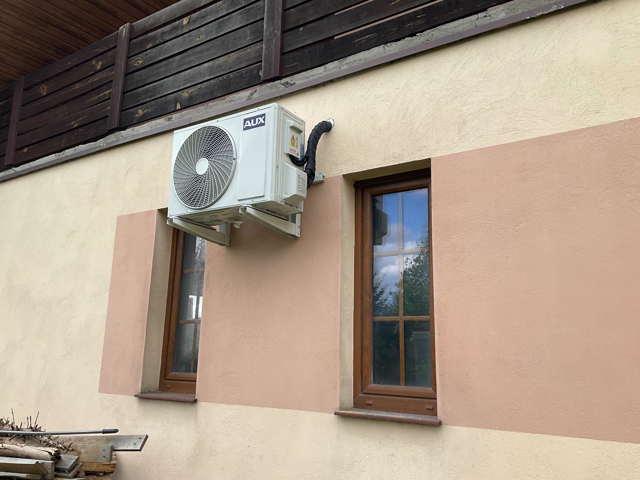 Zewnętrzna jednostka klimatyzatora marki AUX zamontowana na ścianie budynku, widoczne dwa okna z drewnianą ramą i drewniane ogrodzenie w tle.