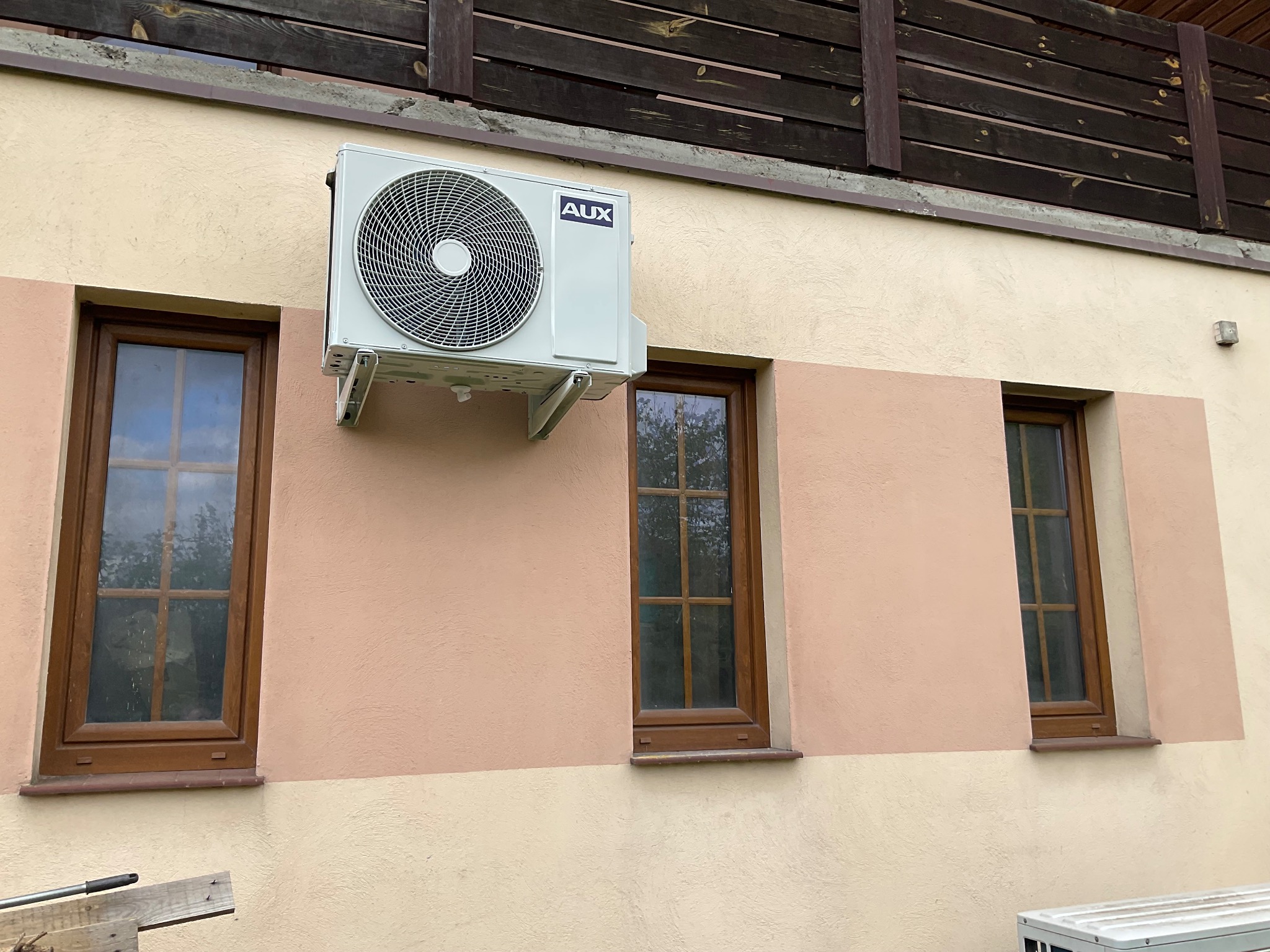 Zewnętrzna jednostka klimatyzatora marki AUX zamontowana na elewacji domu, widoczna pomiędzy oknami z brązowymi ramami, z drewnianą balustradą balkonu w tle.