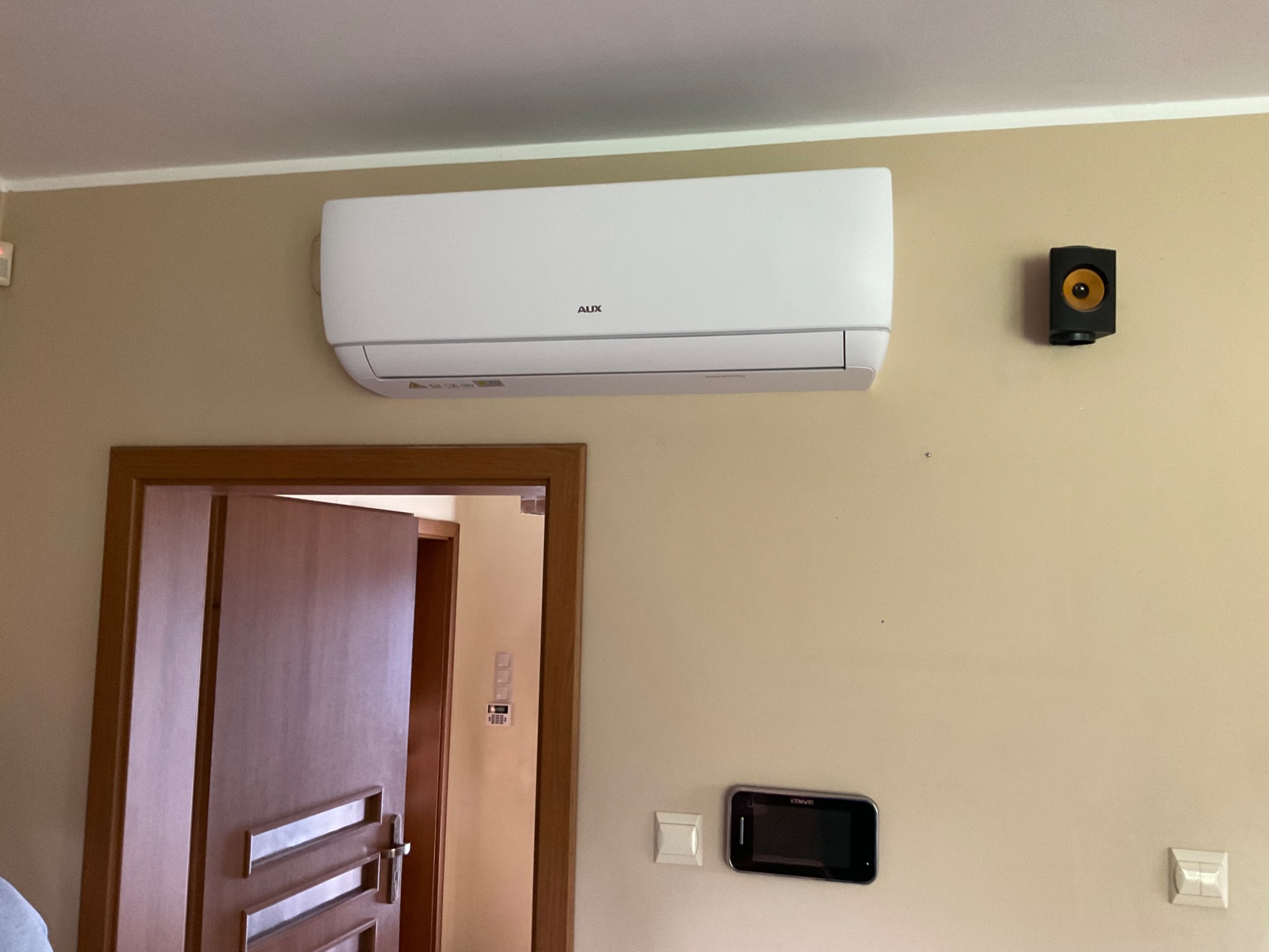 Biały klimatyzator marki AUX zamontowany na beżowej ścianie w domu, obok czarny głośnik i wideodomofon.