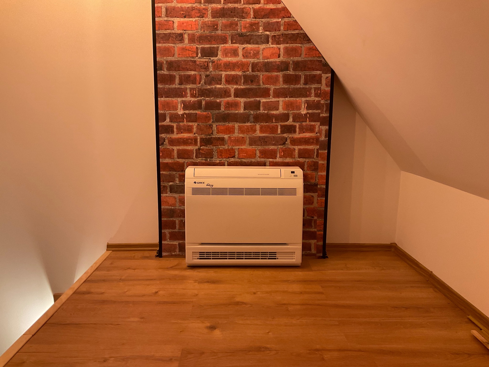 Klimatyzator podłogowy marki Gree ustawiony na drewnianej podłodze na poddaszu, z dekoracyjną ścianą z czerwonej cegły w tle.