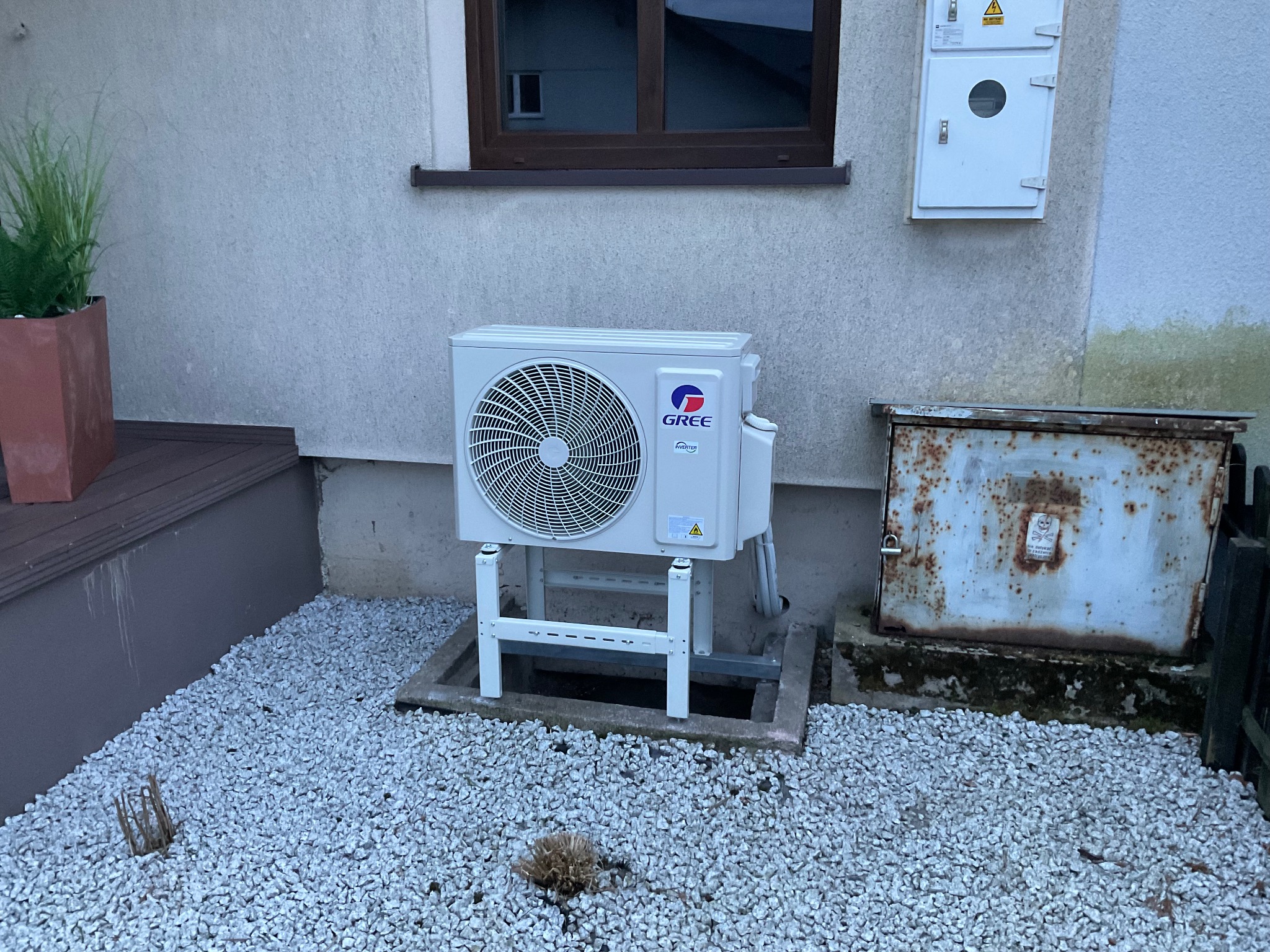Zewnętrzna jednostka klimatyzacji marki Gree, zamontowana na białej metalowej konstrukcji na żwirowym podłożu, obok skorodowanej skrzynki elektrycznej, na tle elewacji domu z oknem.
