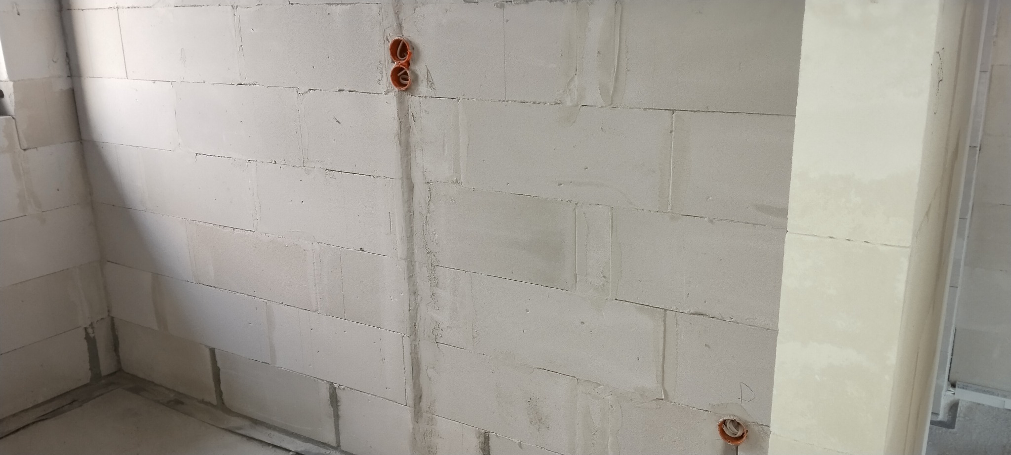 Surowa ściana z betonu komórkowego z widocznymi, pomarańczowymi puszkami instalacyjnymi pod gniazdka elektryczne, przygotowana do dalszych prac wykończeniowych.