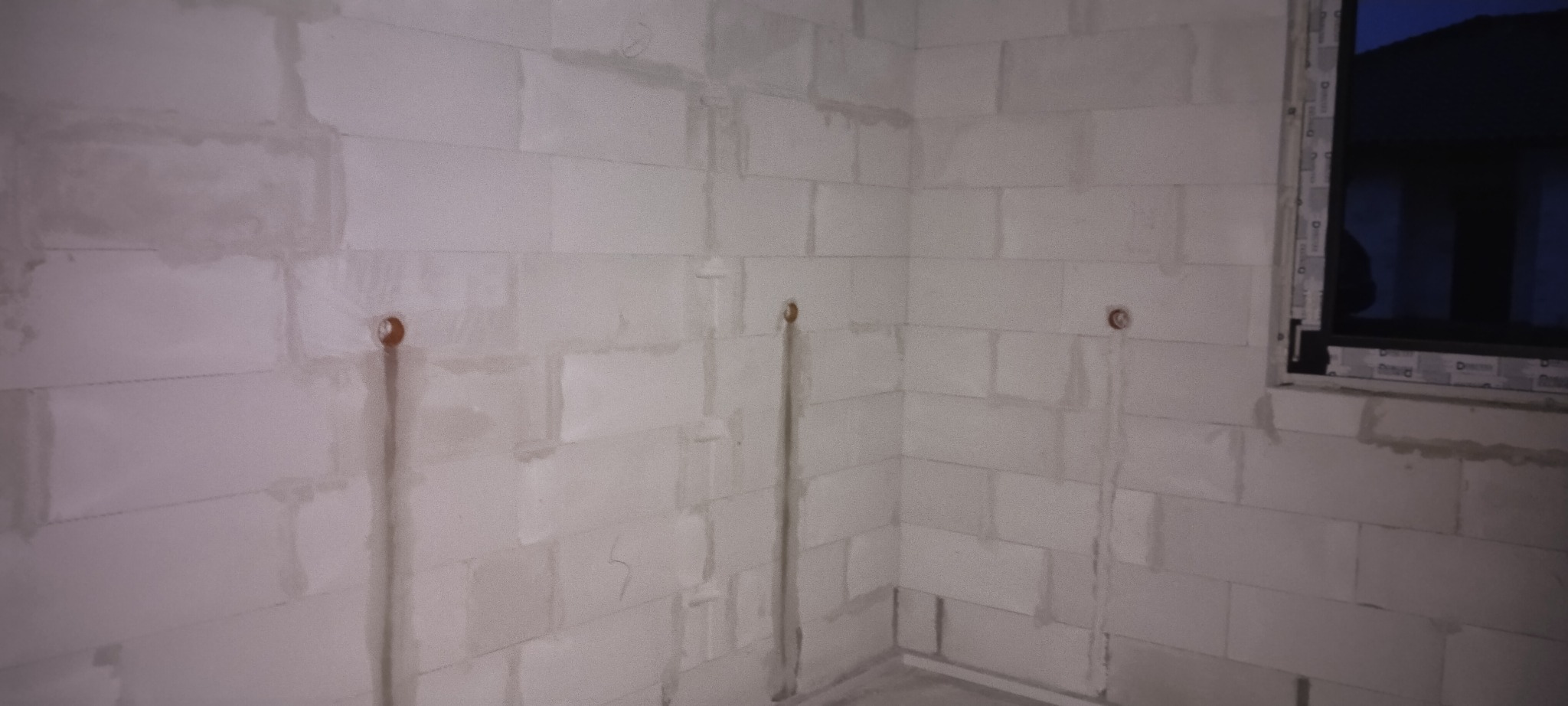 Ściana z betonu komórkowego z widocznymi pionowymi bruzdami pod przewody elektryczne zakończonymi puszkami instalacyjnymi, w tle okno w czarnej ramie.