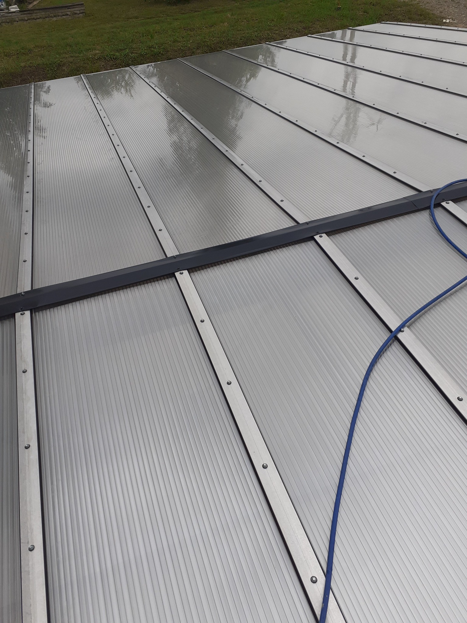 Szary dach z poliwęglanu komorowego z metalowymi profilami i śrubami, częściowo mokry, z fragmentem niebieskiego węża na powierzchni.