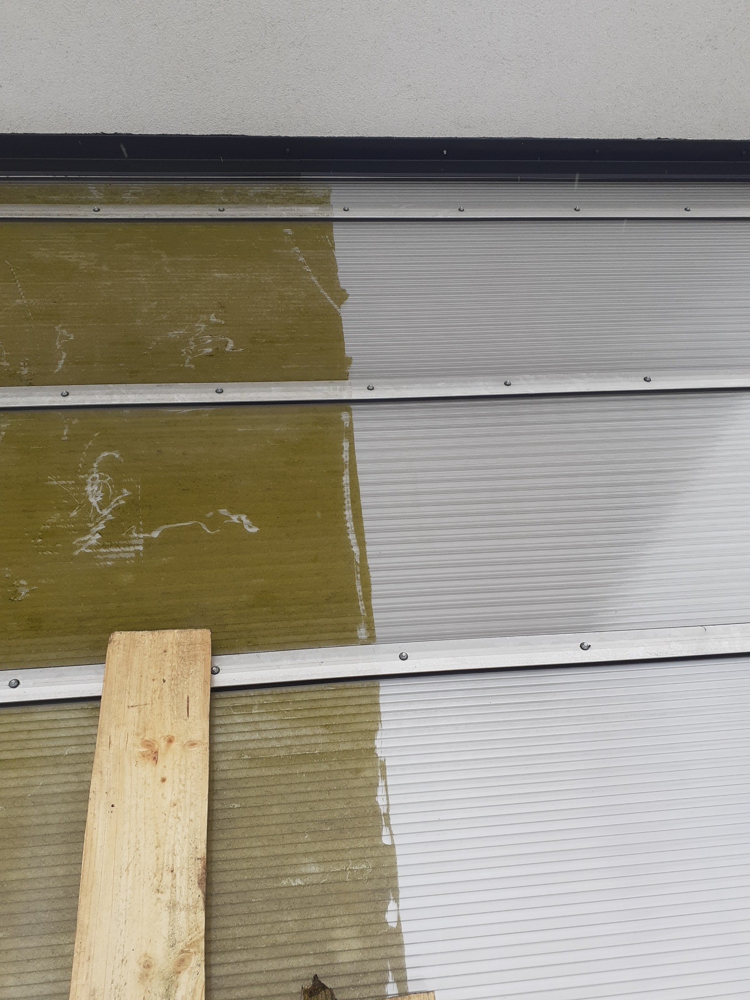 Fragment elewacji z poziomych paneli, w połowie wyczyszczonych, z widoczną różnicą w kolorze i zabrudzeniach, z deską opartą o ścianę.