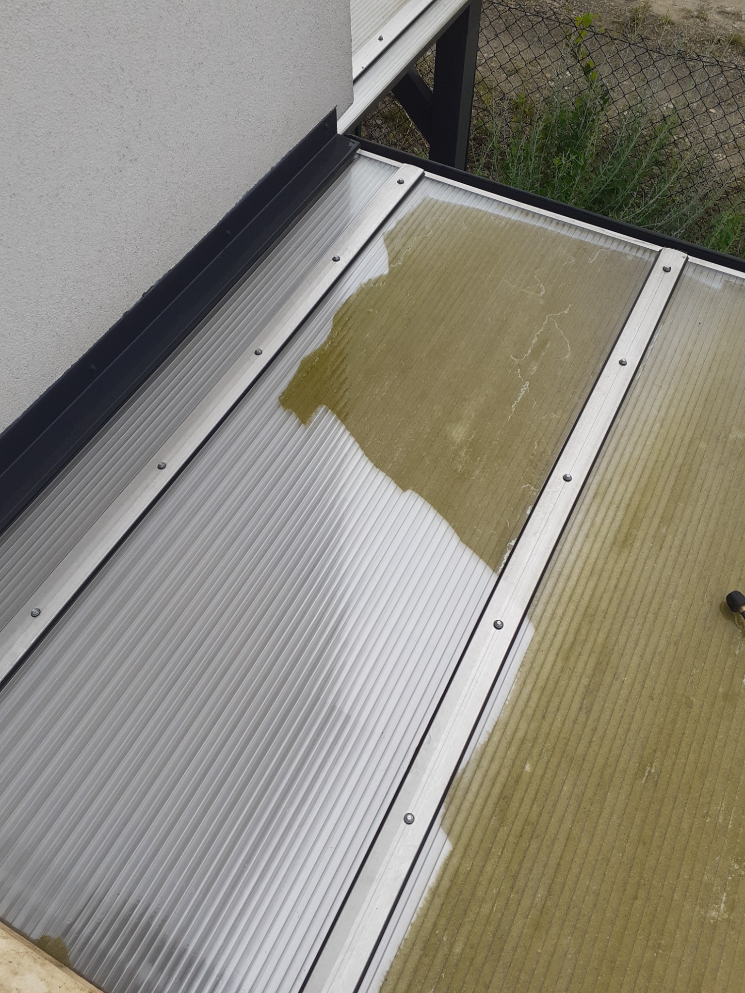 Fragment dachu z poliwęglanu, częściowo umyty, z widoczną różnicą w czystości między umytą a brudną powierzchnią, mocowany aluminiowymi listwami z widocznymi śrubami.