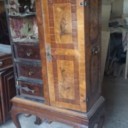 Darek Piotrowski Renowacja mebli , meble na wymiar. - Antyczna, drewniana szafka z otwartymi drzwiami, ukazująca trzy szuflady i uszkodzone wnętrze, stoi na ozdobnych nogach w warsztacie.