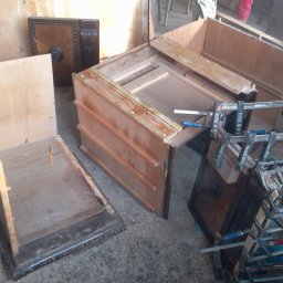 Darek Piotrowski Renowacja mebli , meble na wymiar. - Fragmenty drewnianych mebli w trakcie renowacji, z widocznymi ściskami stolarskimi i elementami dekoracyjnymi, na surowym betonowym podłożu.