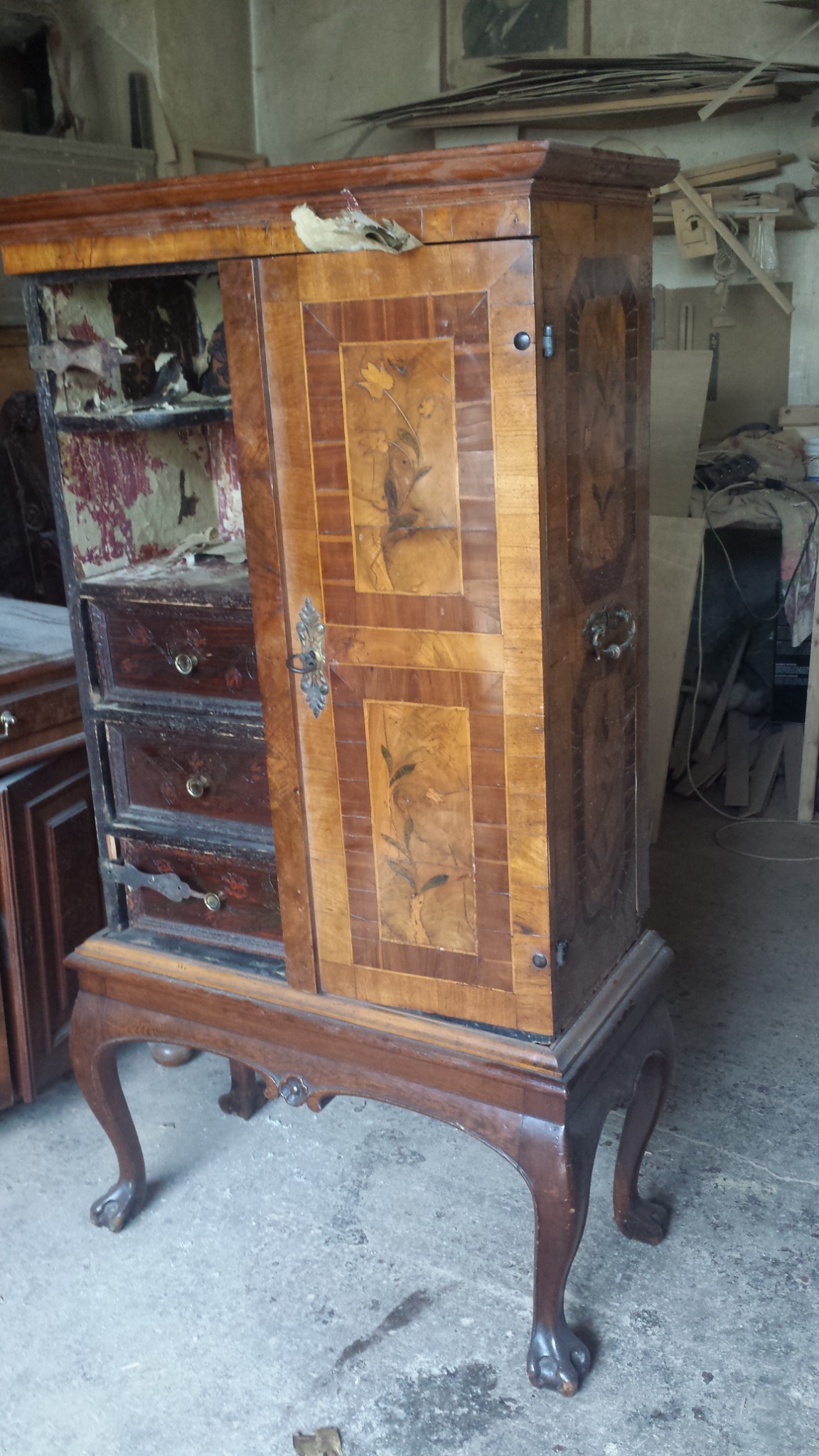 Antyczna, drewniana szafka z otwartymi drzwiami, ukazująca trzy szuflady i uszkodzone wnętrze, stoi na ozdobnych nogach w warsztacie.
