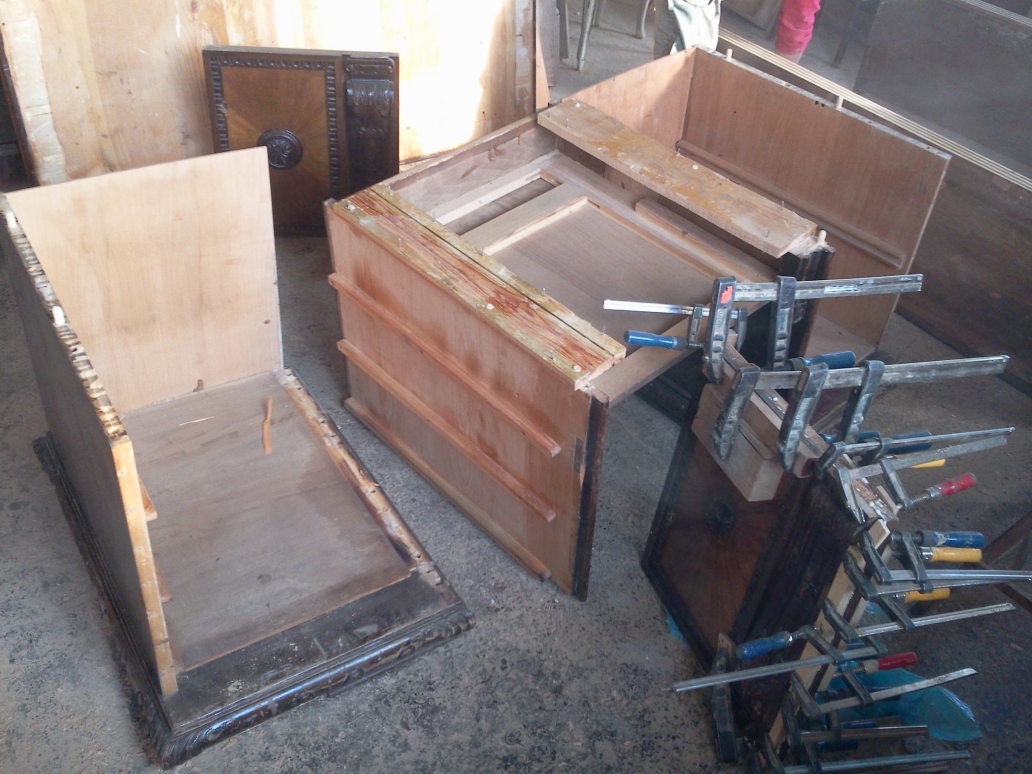 Fragmenty drewnianych mebli w trakcie renowacji, z widocznymi ściskami stolarskimi i elementami dekoracyjnymi, na surowym betonowym podłożu.